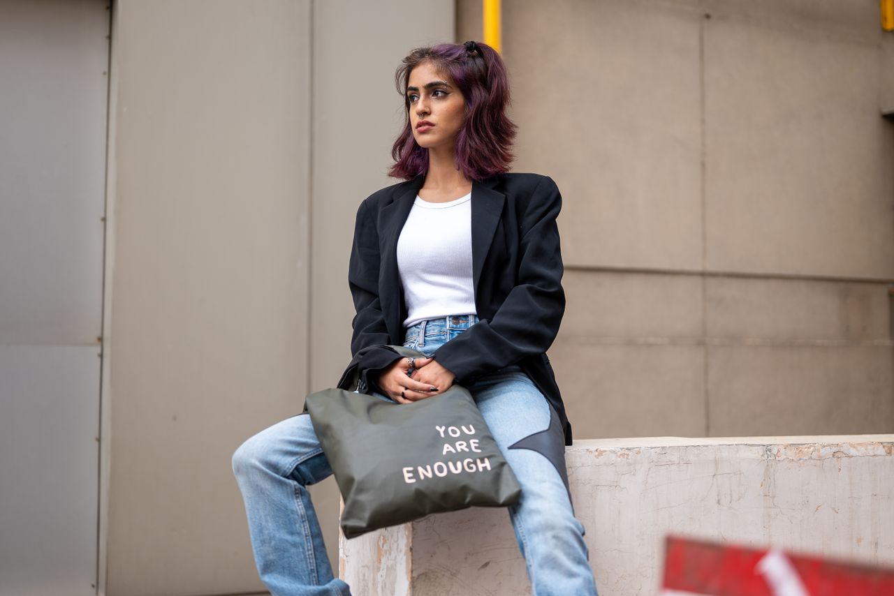 Stylish woman with purple hair sitting outdoors with a motivational tote bag in an urban setting.